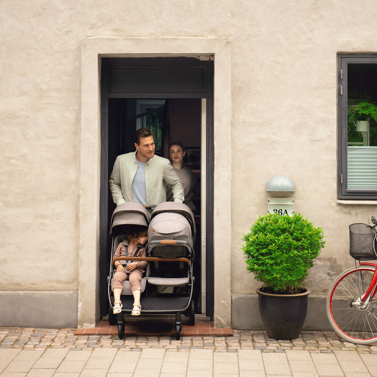 Baby and toddler ride in Nuna TRVL Dubl Stroller - Granite as dad pushes them through doorway