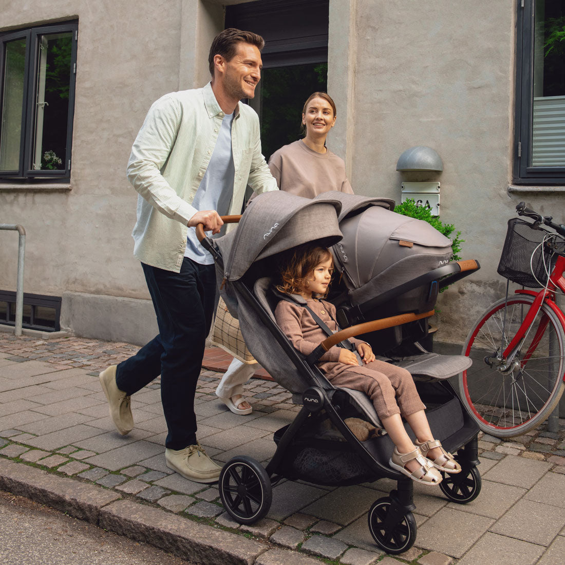 Parents walk down sidewalk while pushing baby in car seat and toddler girl in Nuna TRVL Dubl Stroller - Granite