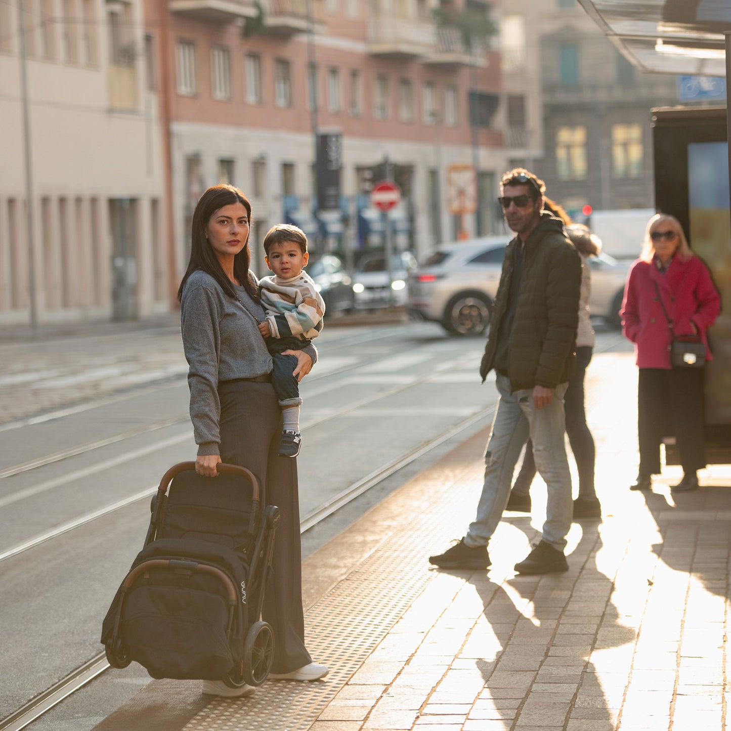 Mom holding folded Nuna TRVL LX Stroller - Caviar next to bus stop