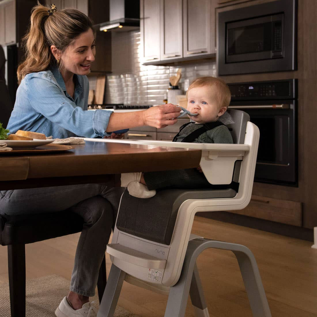 Mom feeds baby sitting in Nuna ZAAZ High Chair with MagneTech Secure Snap - Carbon