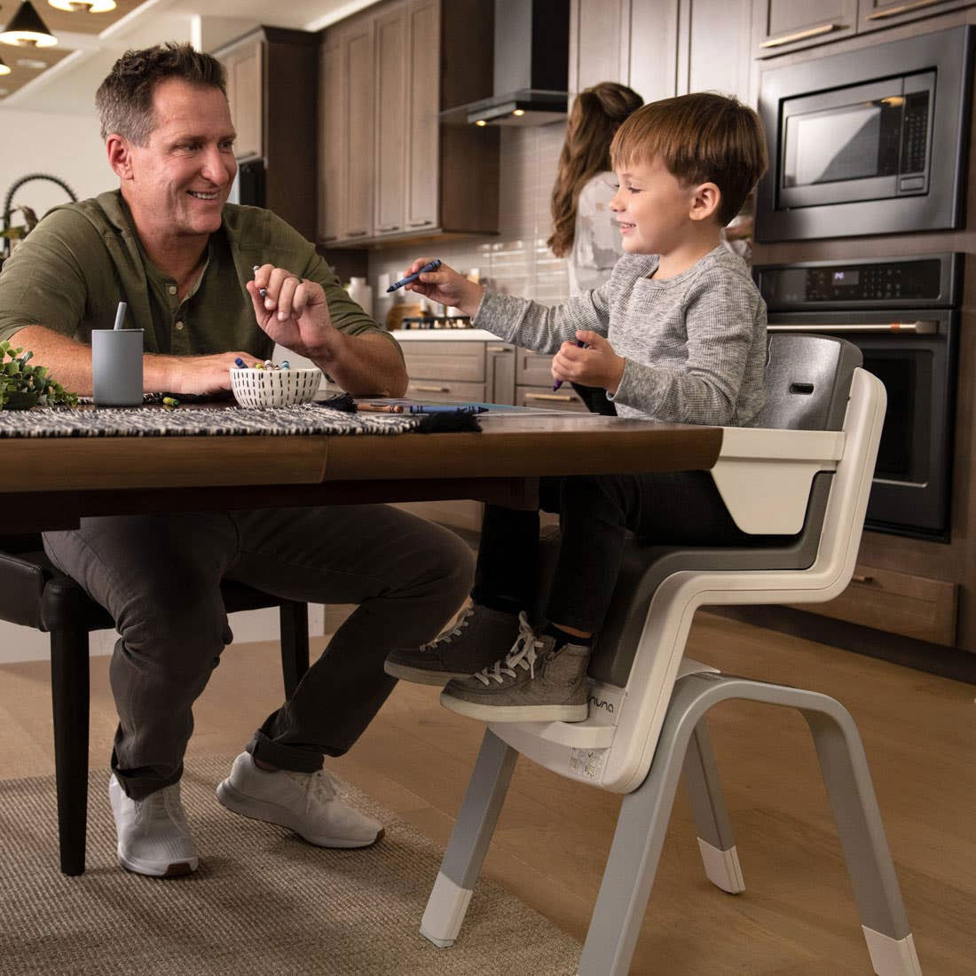 Boy colors at the kitchen table with his father while sitting in Nuna ZAAZ High Chair with MagneTech Secure Snap - Carbon